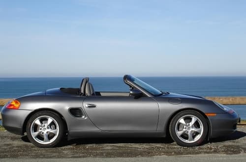 Silbergrauer Porsche Boxster 986 mit offenem Verdeck vor dem Meer. Seitenansicht