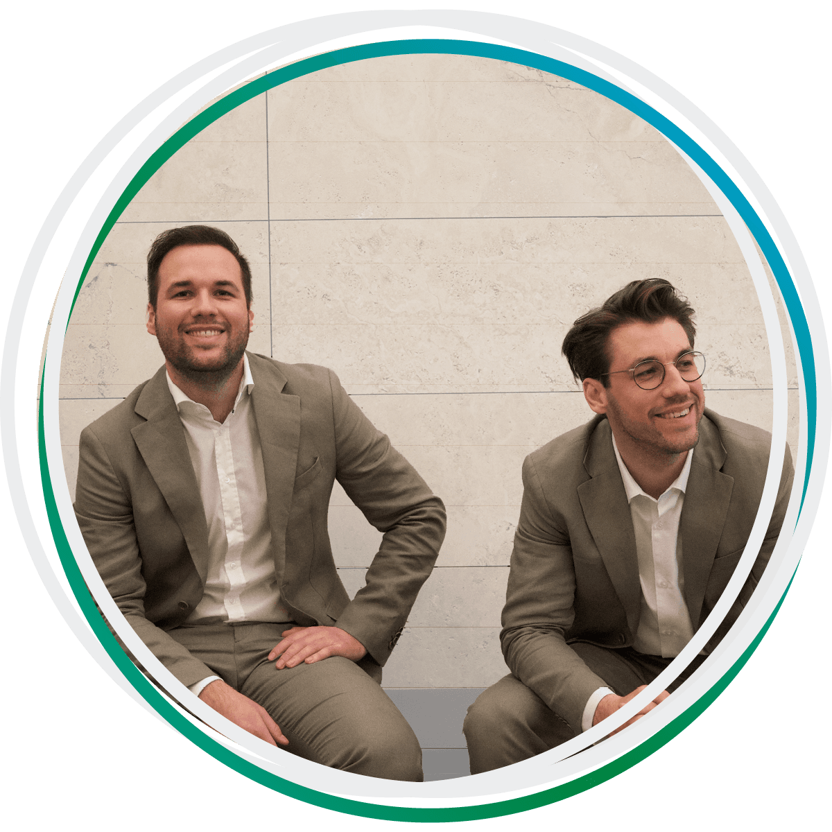 Die Rechtsanwälte Niklas und Lukas Hufeld sitzen vor einer Mauer und lächeln. Beide tragen einen beigen Anzug.