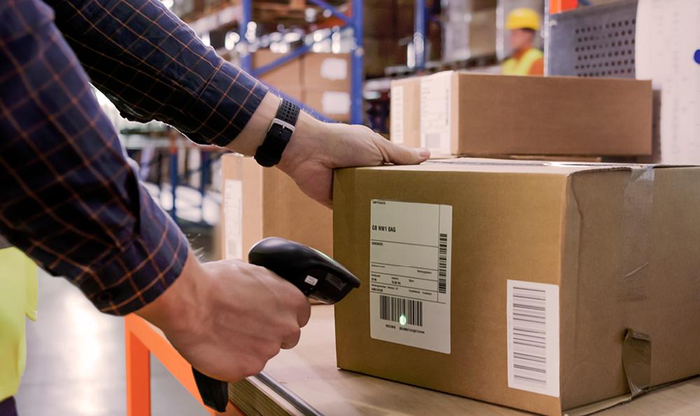 Paket liegt auf einem Tisch in einem Warenlager. Ein Mann in dunkelblauem Hemd hält einen Scanner in der rechten Hand und scannt das Paket en.