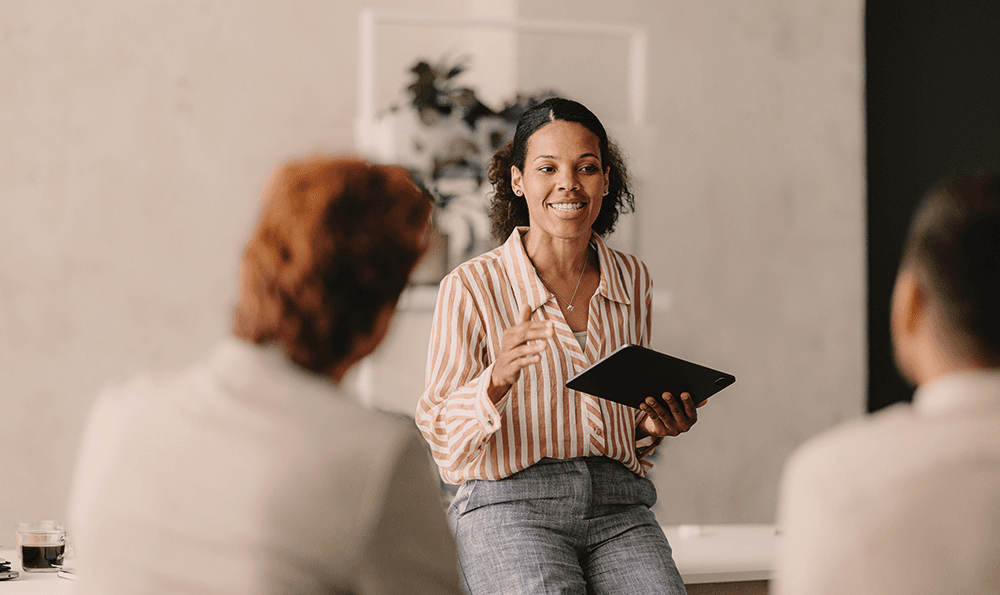 Eine Frau mit einem Tablet unterhält sich mit zwei sitzenden Personen. Sie lächelt, trägt ein gestreiftes Hemd und Jeans, im Hintergrund ist eine Pflanze zu sehen, was eine freundliche und professionelle Atmosphäre vermittelt.