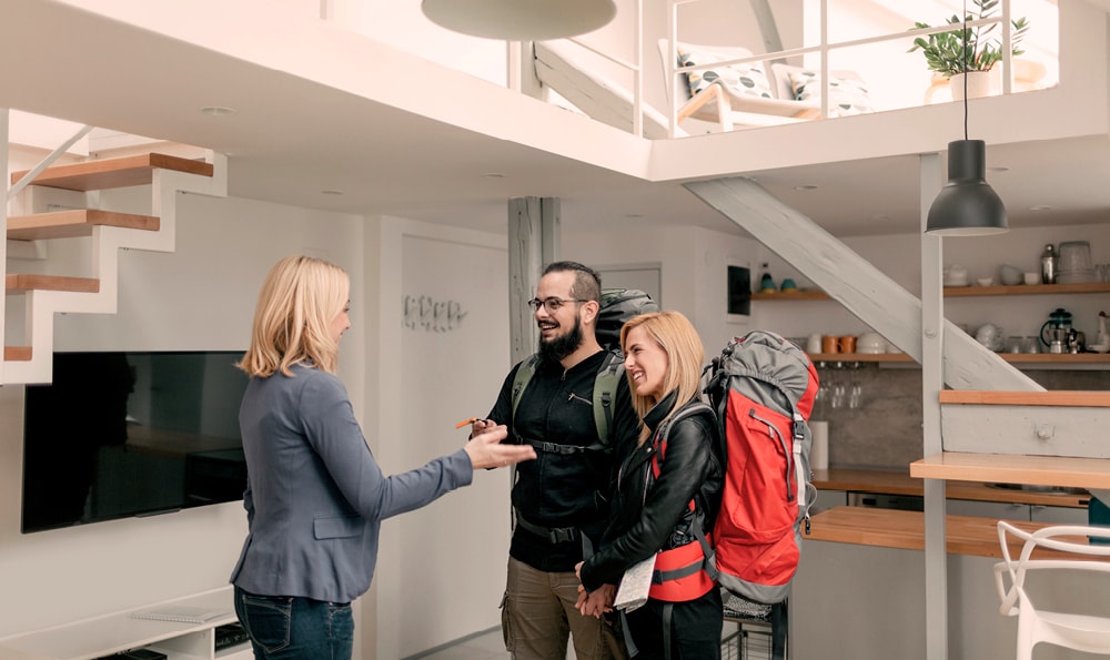 Eine Frau mit blondem Haar übergibt einem lächelnden Paar mit großen Rucksäcken einen Schlüssel für eine moderne Ferienwohnung mit einer Treppe und einem offenen Obergeschoss.