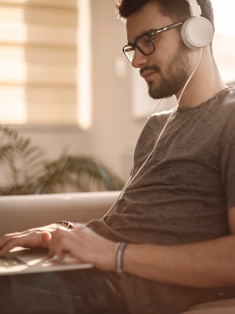 Ein junger Mann mit Brille und Tshirt trägt Kopfhörer und arbeitet an seinem Laptop