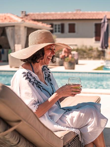 Frau mit breitkrempigem Hut und besticktem Kleid entspannt sich auf einer Liege am Pool, lächelt und hält einen Drink in der Hand. Sonniger Tag in Spanien, lockere und fröhliche Stimmung.