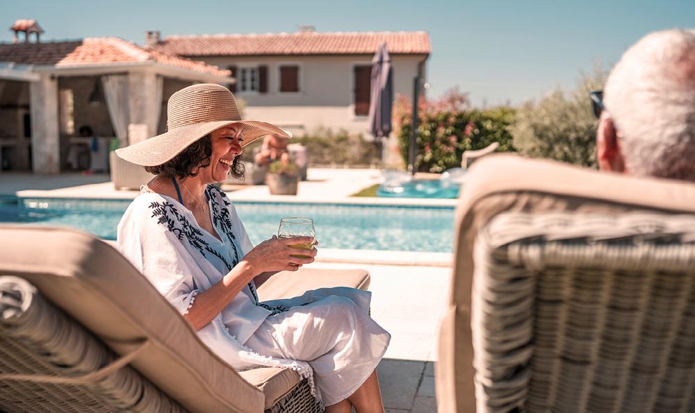 Eine Frau mit einem breitkrempigen Hut lächelt und hält ein Getränk in der Hand, während sie sich am Pool entspannt. Sonniger Tag, entspannte Atmosphäre, mit einem Steinhaus im Hintergrund.
