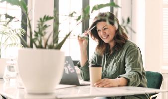 Eine Frau mit langen Haaren sitzt an ihrem Laptop und lächelt. Sie hält einen Kaffeebecher in der Hand und auf dem Tisch steht eine Pflanze