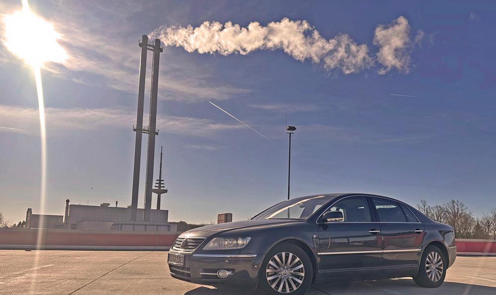 Eine schwarze VW Phaeton D1 3.0 TDI Individual 2007 parkt unter einem klaren blauen Himmel mit einer hellen Sonne in der Ecke. Im Hintergrund steigt Rauch aus hohen Industrieschornsteinen auf.