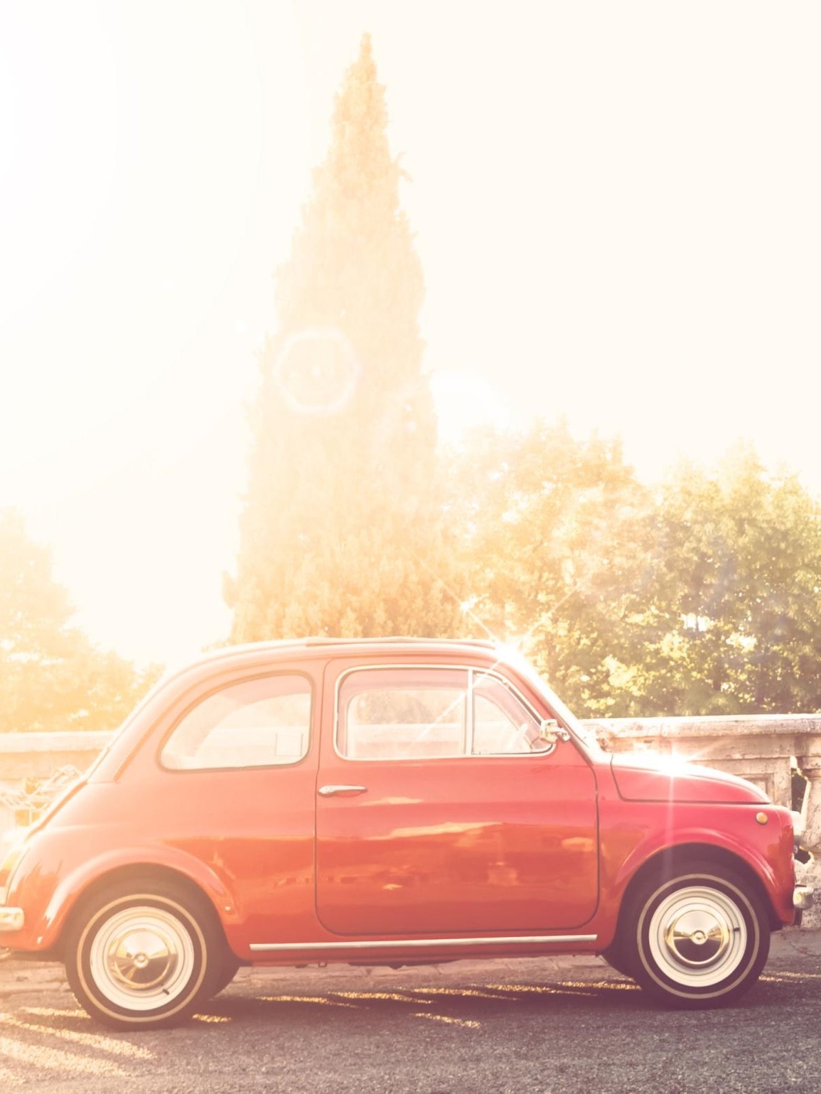Roter Vintage Kleinwagen parkt auf einer Straße neben einer Steingeländermauer, umgeben von Bäumen unter sonnigem Himmel