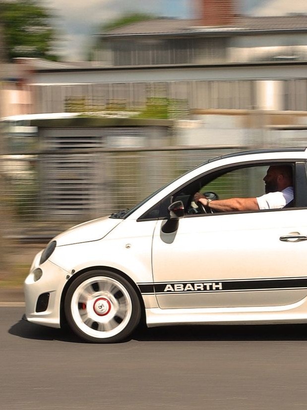 Weißer Abarth 500 mit schwarzen Details ist bei Sonnenschein fahrend auf einer Straße. Man sieht ihn von der Seitenansicht mit männlichem Fahrer hinterm Steuer.