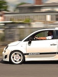 Weißer Abarth 500 mit schwarzen Details ist bei Sonnenschein fahrend auf einer Straße. Man sieht ihn von der Seitenansicht mit männlichem Fahrer hinterm Steuer.