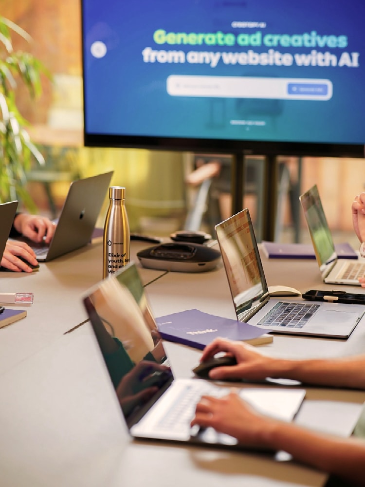 Mehrere Personen arbeiten mit Laptops an einem Konferenztisch. Im Hintergrund zeigt ein Bildschirm eine Präsentation zum Thema KI.