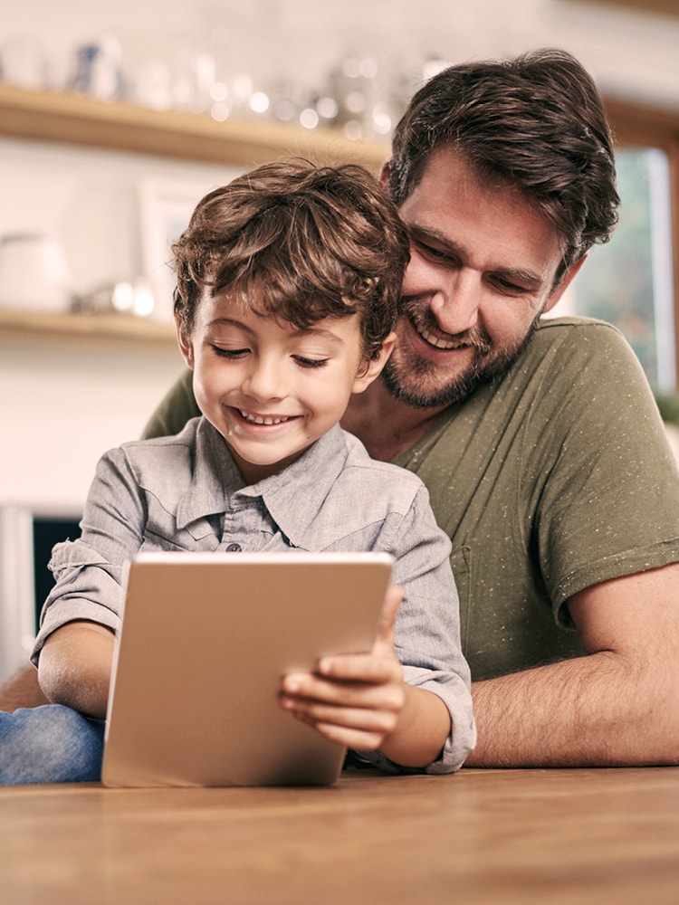 wachsene Person und Kind sitzen zusammen und betrachten ein Tablet in einer Küche.
