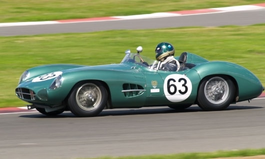 Ein grüner Oldtimer-Rennwagen Aston Martin DBR1 mit der Startnummer 63 rast über die Rennstrecke. Der Fahrer, der einen Helm trägt, beugt sich nach vorne und vermittelt so ein Gefühl von Bewegung und Spannung.