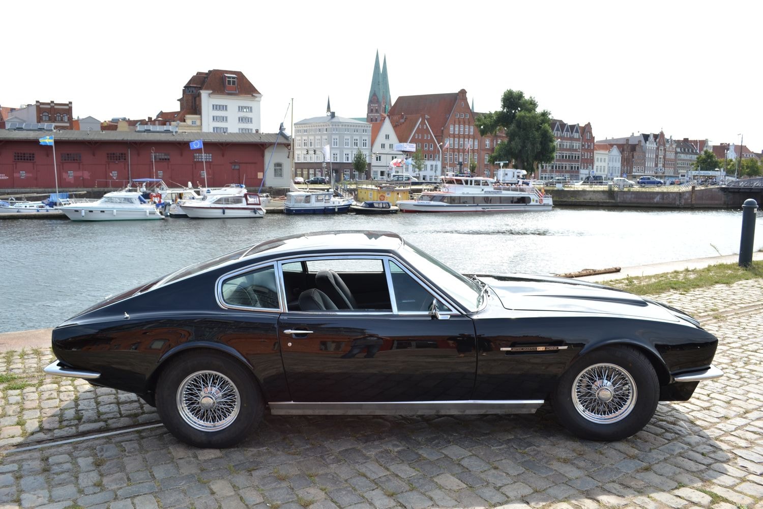 Schwarzer Aston Martin geparkt an einem Flussufer mit historischen Gebäuden und Booten im Hintergrund