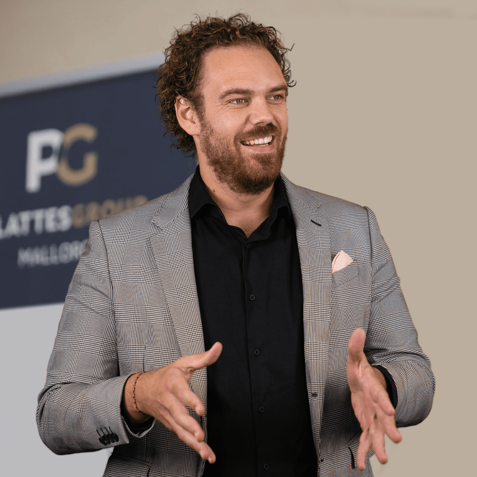 Christian Plattes mit lockigem Haar und Bart, in einem grauen Anzug und schwarzem Hemd, lächelnd und gestikulierend. Im Hintergrund ist ein unscharfes Schild mit dem „PG“-Logo zu sehen.