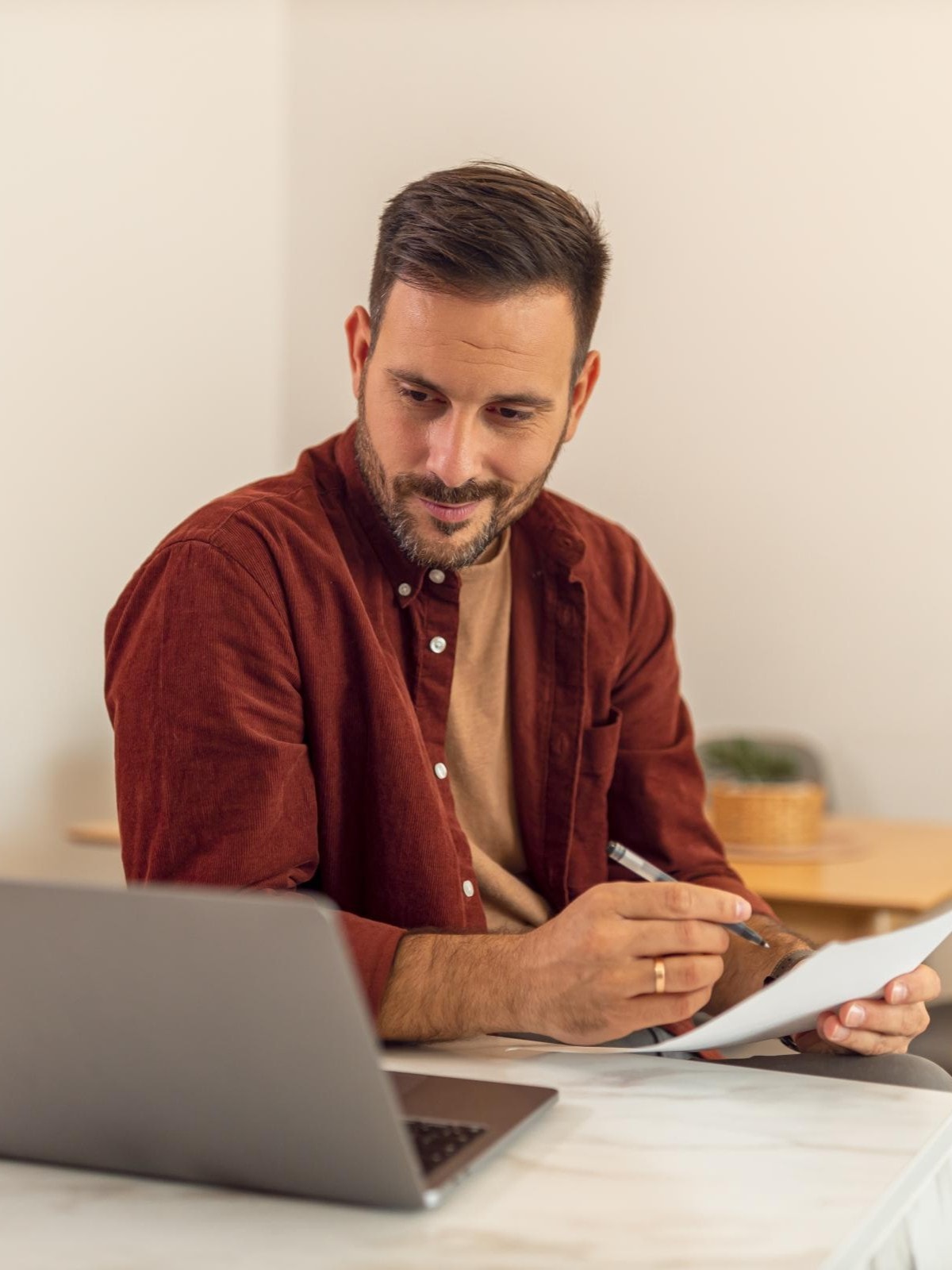 Ein Mann im mittleren Alter mit braunen Haaren und dunkelrotem Hemd sitzt vor seinem Laptop am Tisch und macht sich Notizen. Neben dem Laptop liegt ein Taschenrechner.