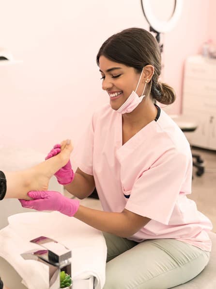 Junge Fußpflegerin in rosa Arbeitskleidung sitzt in ihrem Studio auf einem Hocker und behandelt die Füße einer Kundin.