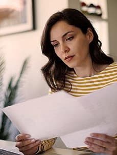 Junge Frau mit braunen Haaren und gestreiftem Oberteil sitzt an einem Schreibtisch vor ihrem Laptop. In den Händen hält sie Geschäftsunterlagen, die sie für ihre Steuererklärung als Selbstständige benötigt.