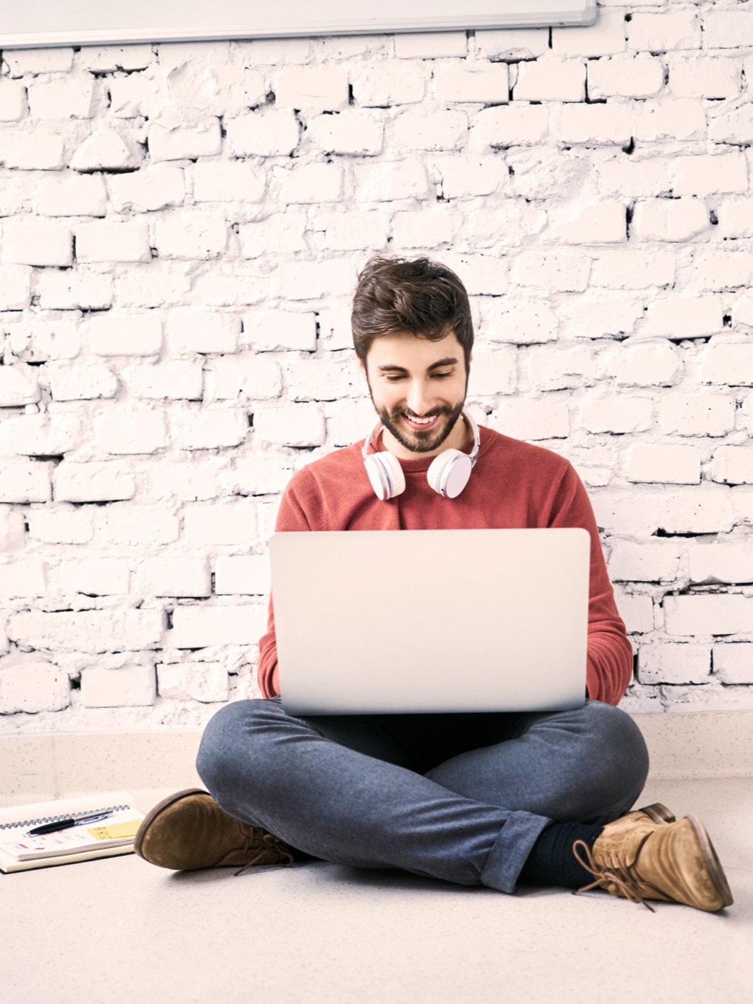 Junger Mann mit Bart und Kopfhörern um den Hals sitzt lächelnd am Boden vor einer weißen Ziegelwand und arbeitet an einem Laptop
