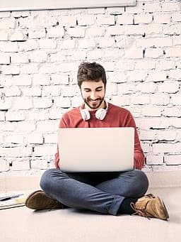 Junger Mann mit Bart und Kopfhörern um den Hals sitzt lächelnd am Boden vor einer weißen Ziegelwand und arbeitet an einem Laptop