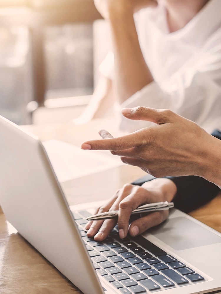 Drei Menschen, von denen jeweils die Hände zu sehen sind, sitzen an einem Schreibtisch, auf dem sich ein Laptop und Unterlagen befinden, und diskutieren miteinander