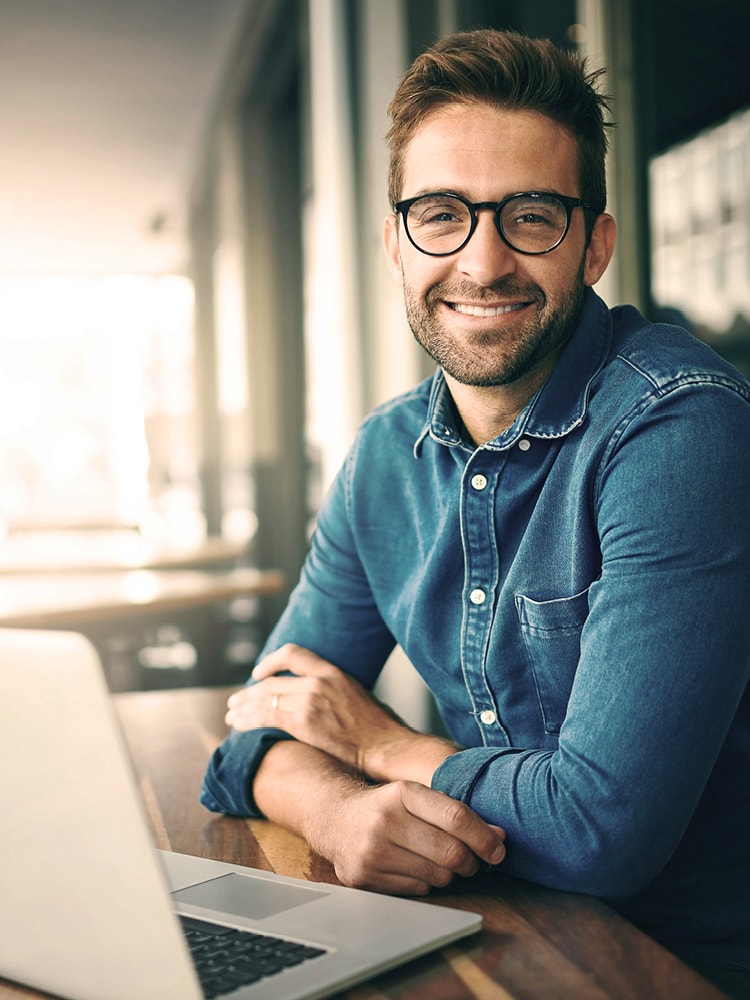 Ein Mann in Hemd und mit Brille sitzt vor seinem Laptop und lächelt in die Kamera