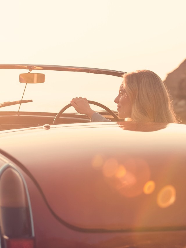 Eine Frau am Steuer eines roten Oldtimers mit offenem Verdeck während einer Fahrt bei tief stehender Sonne.