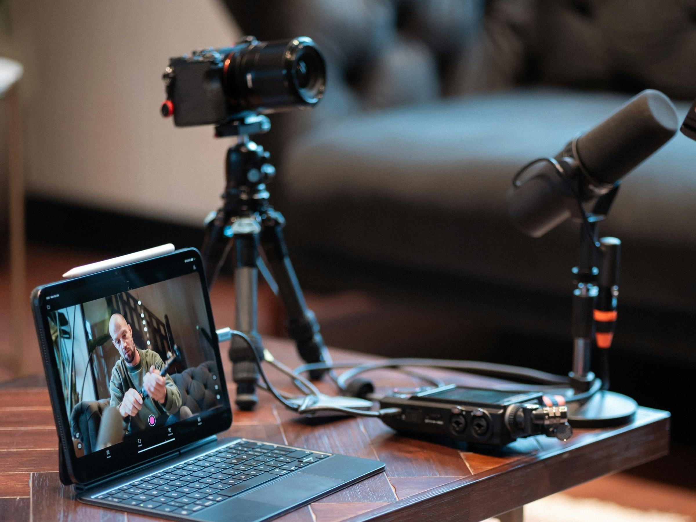 Ein Podcast-Setup zu Hause auf einem Holztisch, bestehend aus einem Laptop, auf dem ein Interview zu sehen ist, einer Kamera auf einem Stativ, Mikrofonen und Audiogeräten. Gemütliche Atmosphäre.