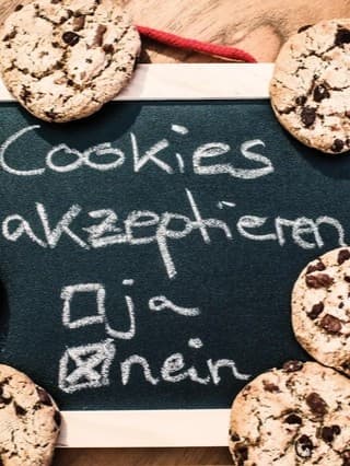 Eine kleine Tafel, umgeben von Schokoladenkeksen, mit der Aufschrift "Cookies akzeptieren". Darunter sind zwei Optionen: "ja" mit einem leeren Kontrollkästchen und "nein" mit einem angekreuzten Kontrollkästchen. Rechts von der Tafel liegt ein Notizblock mit dem Wort "DATENSCHUTZ"