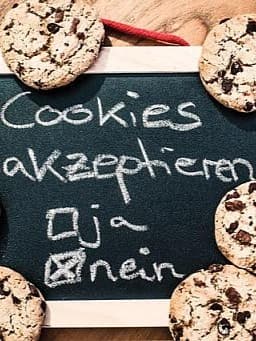 Eine kleine Tafel, umgeben von Schokoladenkeksen, mit der Aufschrift "Cookies akzeptieren". Darunter sind zwei Optionen: "ja" mit einem leeren Kontrollkästchen und "nein" mit einem angekreuzten Kontrollkästchen. Rechts von der Tafel liegt ein Notizblock mit dem Wort "DATENSCHUTZ"