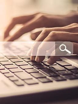 Im Hintergrund sind zwei Hände, die auf einem Laptop tippen und im Vordergrund ist eine Illustration von einem Suchfeld mit einer Lupe.