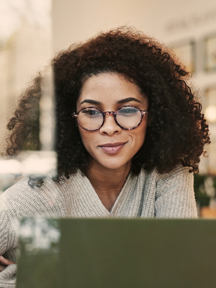 Eine Frau mit Locken und eine Brille blickt lächelnd auf den Bildschirm Ihres Laptops