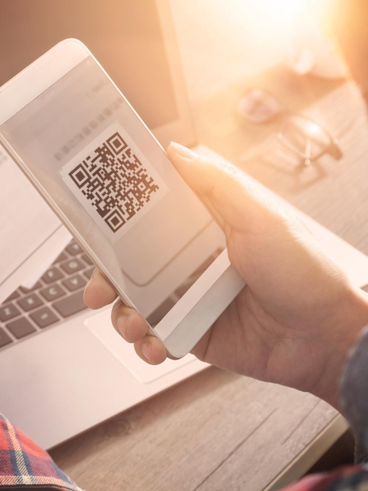 Person scannt mit einem Smartphone einen QR-Code auf einem Papierdokument, im Hintergrund sind ein Laptop und eine Brille sichtbar