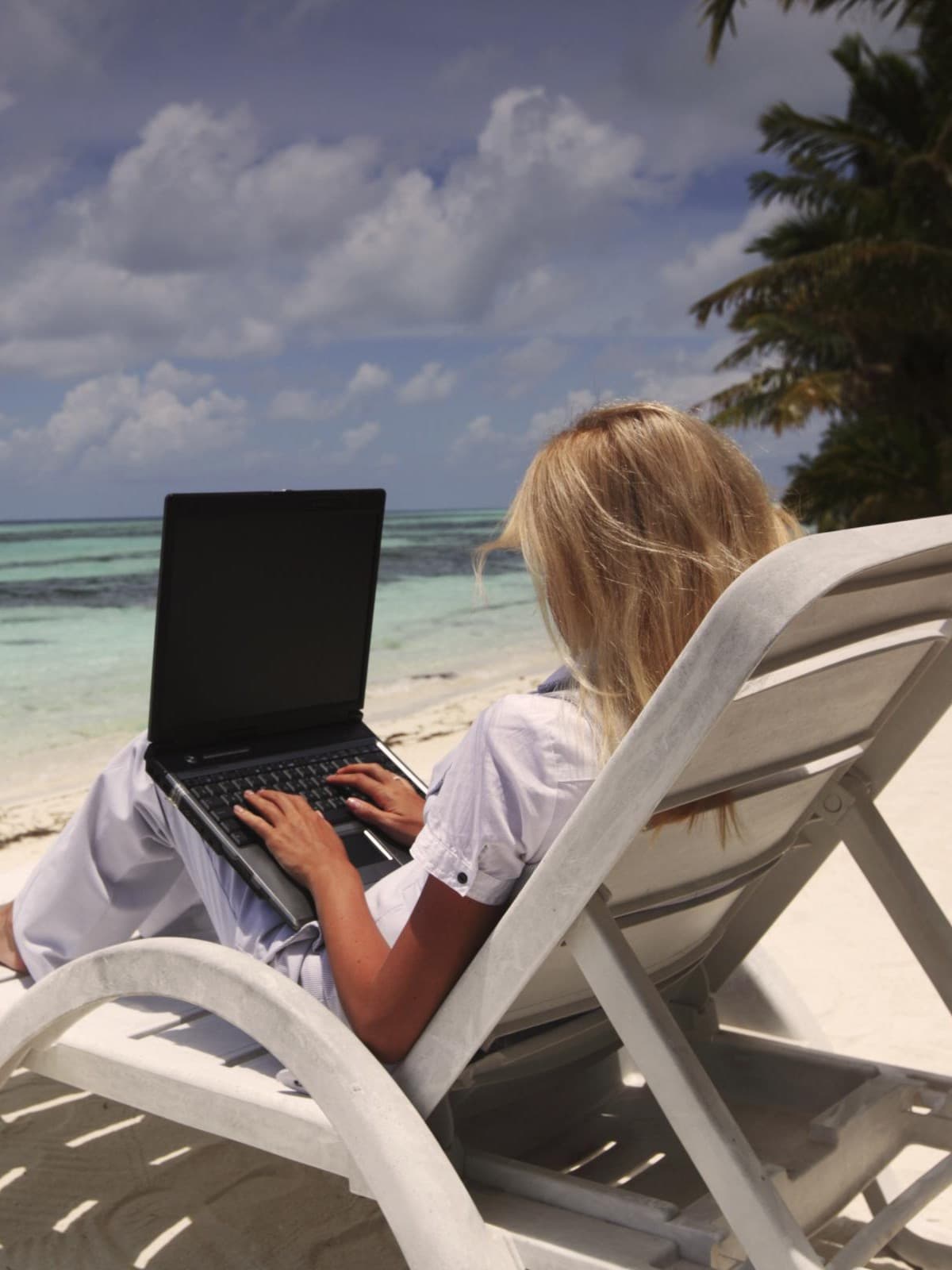 Frau sitzt auf einer Liege am Strand und arbeitet an einem Laptop, im Hintergrund das türkisfarbene Meer und Palmen