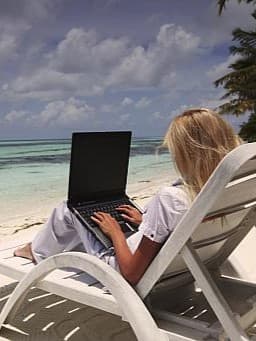 Frau sitzt auf einer Liege am Strand und arbeitet an einem Laptop, im Hintergrund das türkisfarbene Meer und Palmen