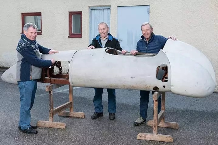 Drei ältere Männer stehen draussen und halten gemeinsam eine unfertige Karosserie eines Oldtimers, die auf Holzgestellen abgestützt ist.