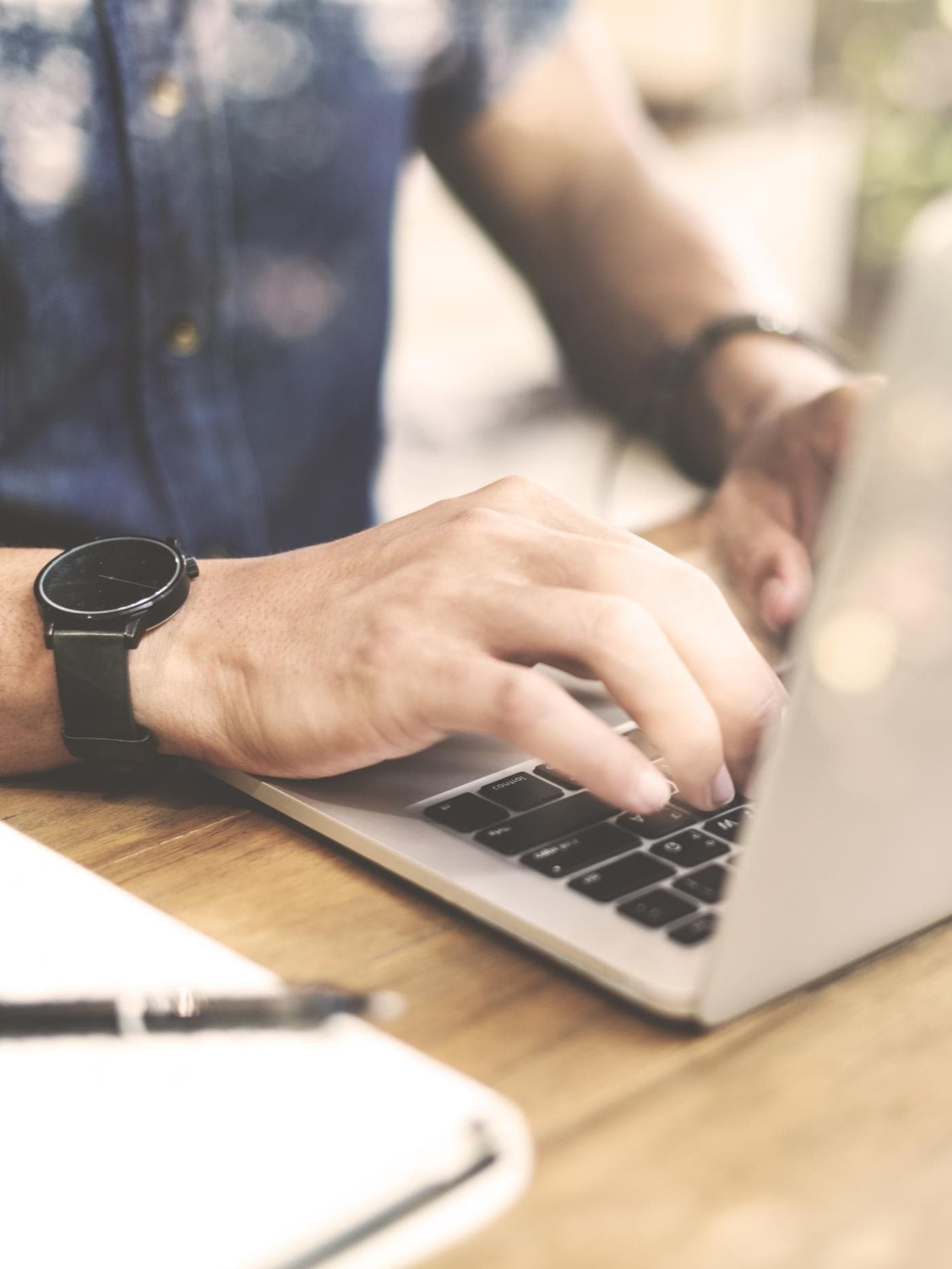Eine Person, die ein blaues Hemd und eine schwarze Uhr trägt, tipp auf einen Laptop. Das Gesicht der Person ist nicht zu sehen.