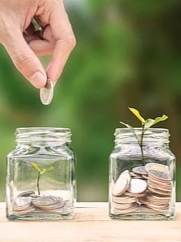 Eine Hand legt Münzen in drei Gläser, in denen Pflanzen wachsen, symbolisiert Investition in nachhaltiges Wachstum.