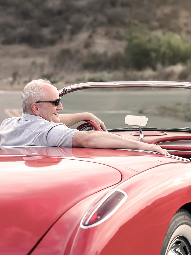 Ein Mann in Hemd und Sonnenbrille sitzt entspannt in einem roten Cabrio-Oldtimer