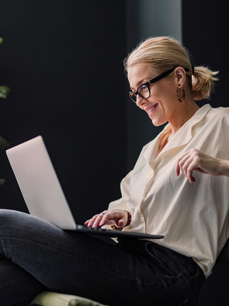 Eine Frau mit Brille und weißer Bluse sitzt auf einem Stuhl und blickt lächeln auf den Bildschirm ihres Laptops, der auf ihrem Schoß ist