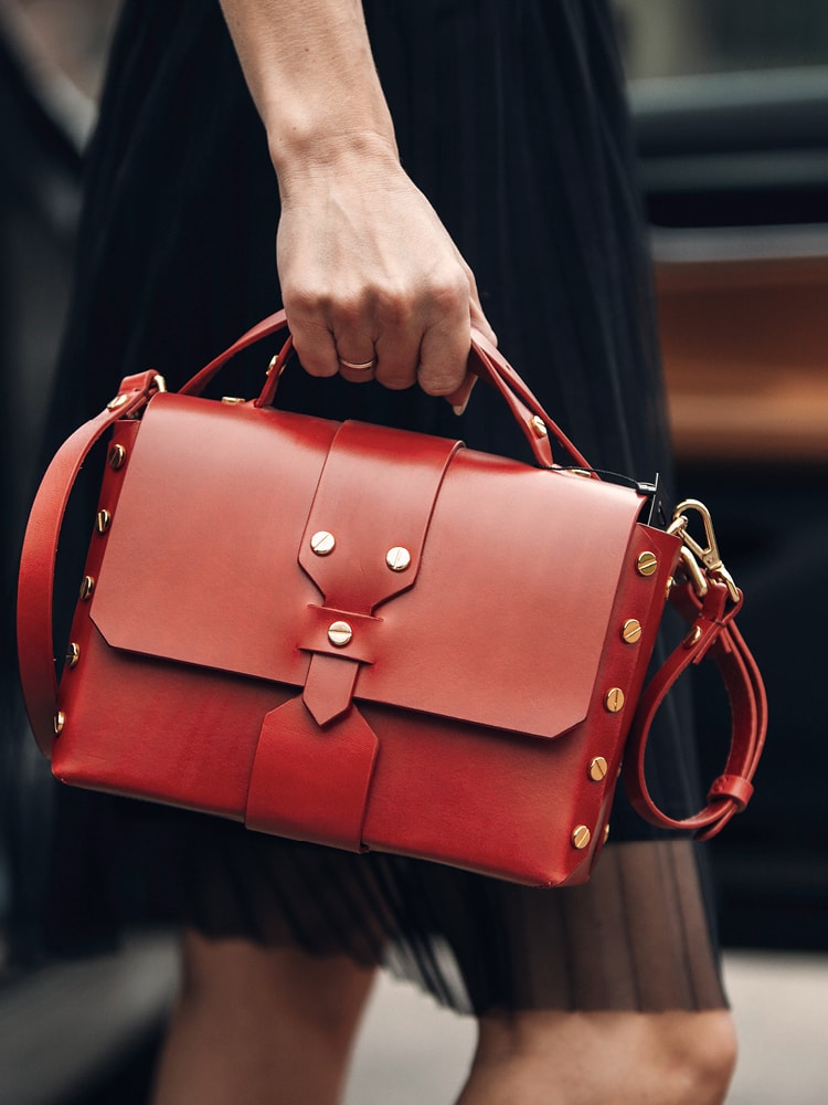 Eine Frau in einem Kleid befindet sich vor einem Auto und hält eine rote Handtasche mit Nieten in der Hand.