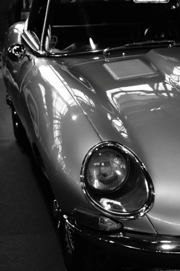 Schwarz-Weiß-Bild eines Oldtimer-Sportwagens Jaguar E-Type mit Fokus auf die vordere rechte Seite. Reflektierende Kurven und runde Scheinwerfer vermitteln einen klassischen, eleganten Stil.