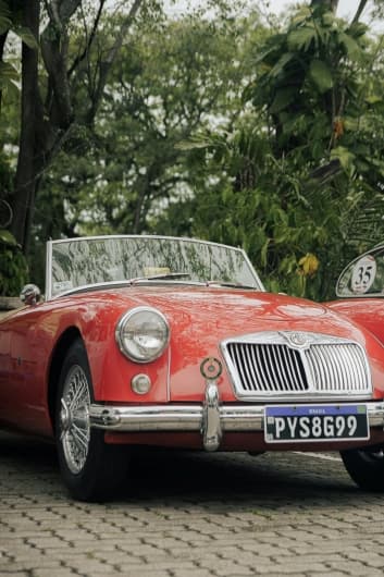 Ein klassisches rotes MG-A Cabrio mit glänzendem Chromgrill und Speichenrädern steht auf einem Kopfsteinpflasterweg, umgeben von üppigem Grün und Bäumen.