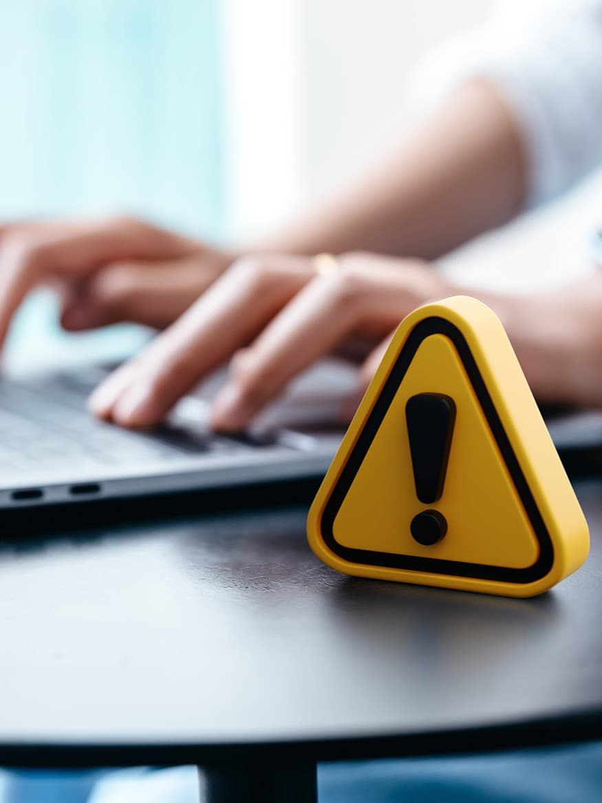 Person, die an einem Tisch auf einem Laptop tippt, mit einem gelben Warndreieck mit Ausrufezeichen im Vordergrund, das zur Vorsicht oder Wachsamkeit mahnt.