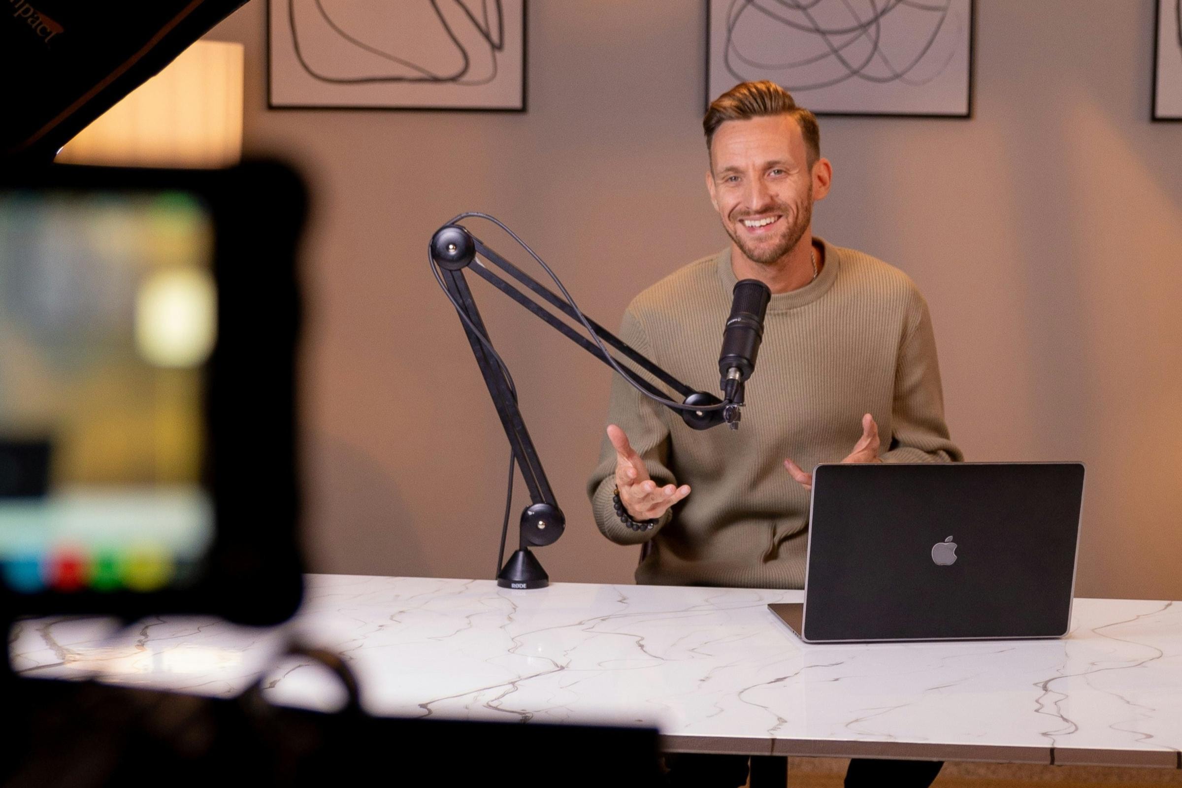 Ein Mann, der während einer Podcast-Aufnahme lächelt und in ein Mikrofon spricht. Er sitzt an einem Marmortisch mit einem Laptop, im Hintergrund sind gerahmte Bilder zu sehen.