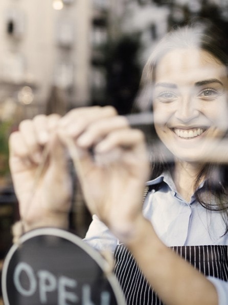 Eine lächelnde Frau in einer gestreiften Schürze öffnet ein Café und richtet ein Schild an der Glastür aus. Die Szene ist warm und einladend, mit verschwommener Straßenansicht draußen.
