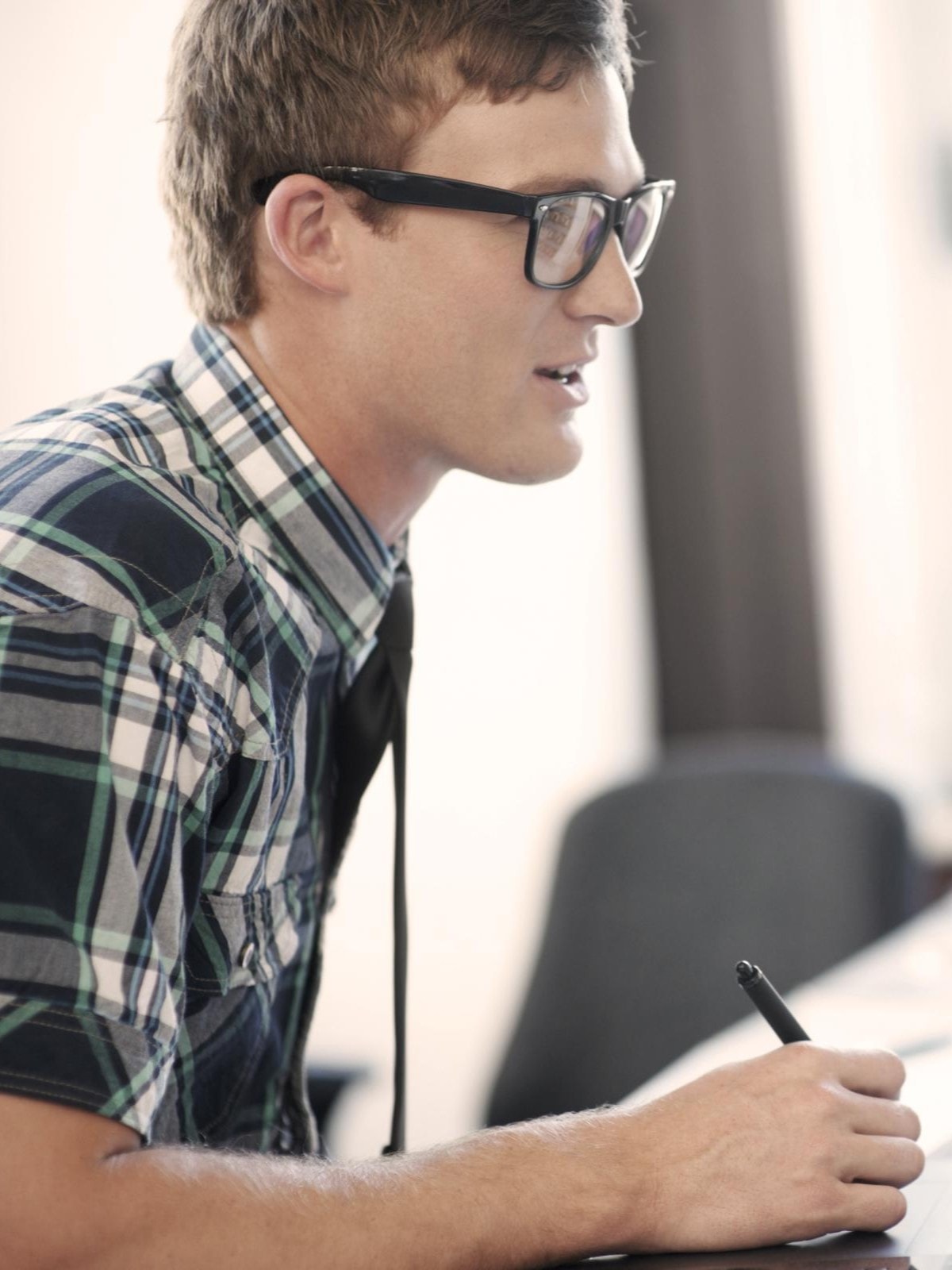 Das Bild zeigt eine Person, die an einem Schreibtisch sitzt und an einem Computer arbeitet. Die Person hält einen Stift und scheint auf einem Tablet oder Papier vor dem Computer zu schreiben oder zu zeichnen. Der Arbeitsplatz umfasst mehrere Monitore, Papiere und Büromaterialien