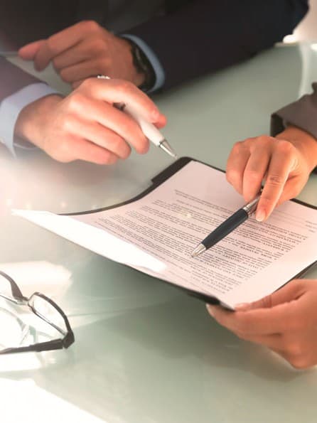 Hände von einer Frau und einem Mann in Business-Kleidung, die an einem Tisch einen Vertrag besprechen; eine Brille liegt daneben.