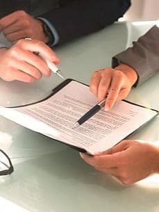 Hände von einer Frau und einem Mann in Business-Kleidung, die an einem Tisch einen Vertrag besprechen; eine Brille liegt daneben.