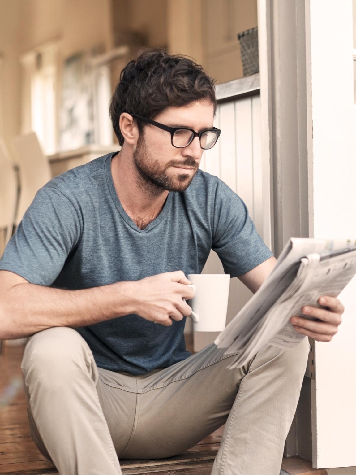 Ein Mann sitzt auf den Stufen seines Hauses, liest eine Zeitung und hält dabei eine Tasse in der Hand.