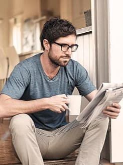 Ein Mann sitzt auf den Stufen seines Hauses, liest eine Zeitung und hält dabei eine Tasse in der Hand.
